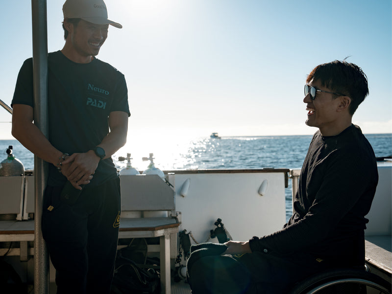 Neuro founders on a boat with one in a wheelchair, smiling and looking at each other, with the ocean in the background.