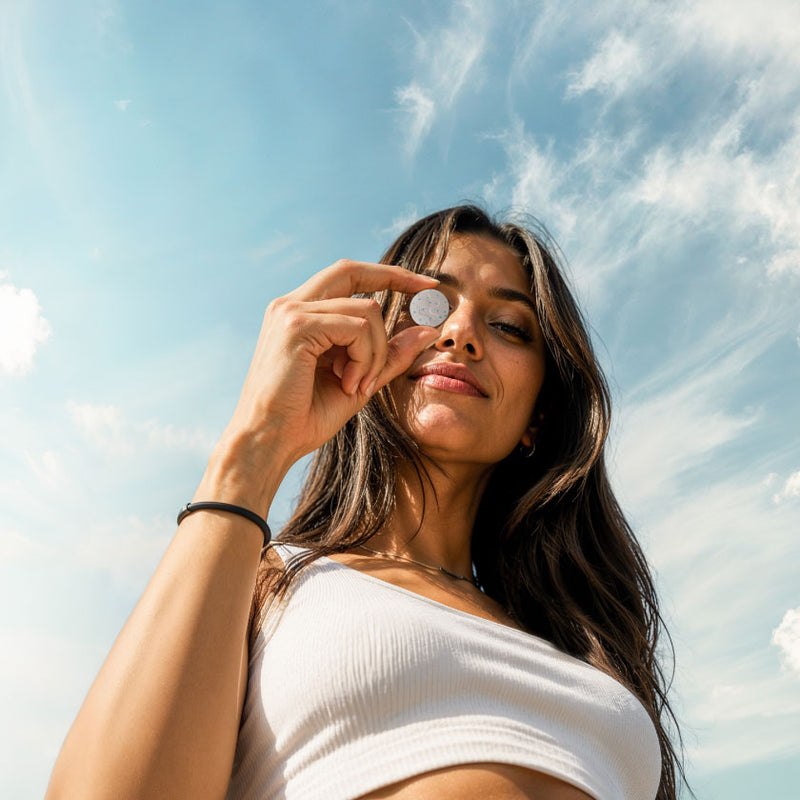 A woman with long dark hair wearing a white ribbed off-the-shoulder crop top, holding Neuro product up to her eye against a bright blue sky with wispy white clouds.