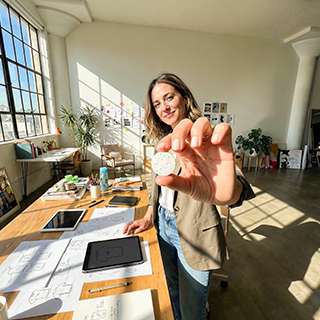 A woman in a bright, sunlit studio holds up a small round white object toward the camera, with a desk full of papers, tablets, and design tools in the background.