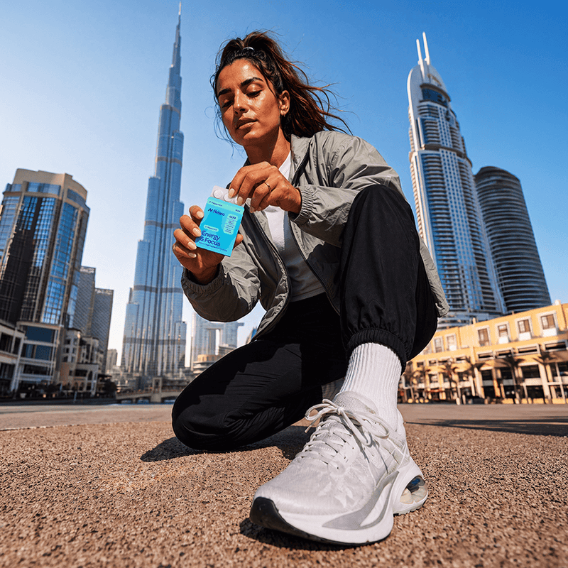 A woman crouches on a city plaza with skyscrapers in the background, holding a small blue drink pouch with a straw. She wears a light gray jacket, black pants, white socks, and white sneakers.