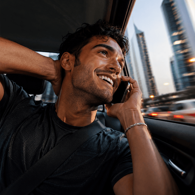 A smiling man in a black t-shirt sitting in a car, talking on a mobile phone. The background shows blurred city skyscrapers and street lights at dusk.