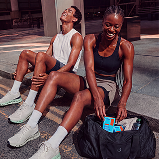 Two athletes sitting on outdoor concrete steps beside a black duffel bag. The woman in a black sports bra and gray shorts is packing items into the bag, while the man in a white sleeveless shirt and dark shorts sits beside her wearing white running shoes.