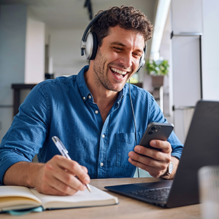 A man wearing over-ear headphones sits at a desk, smiling while holding a smartphone and writing in a notebook. A laptop is open in front of him in a bright indoor setting.