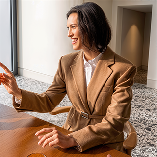 A woman wearing a camel brown belted blazer over a white collared shirt, seated at a wooden table in a modern indoor setting.
